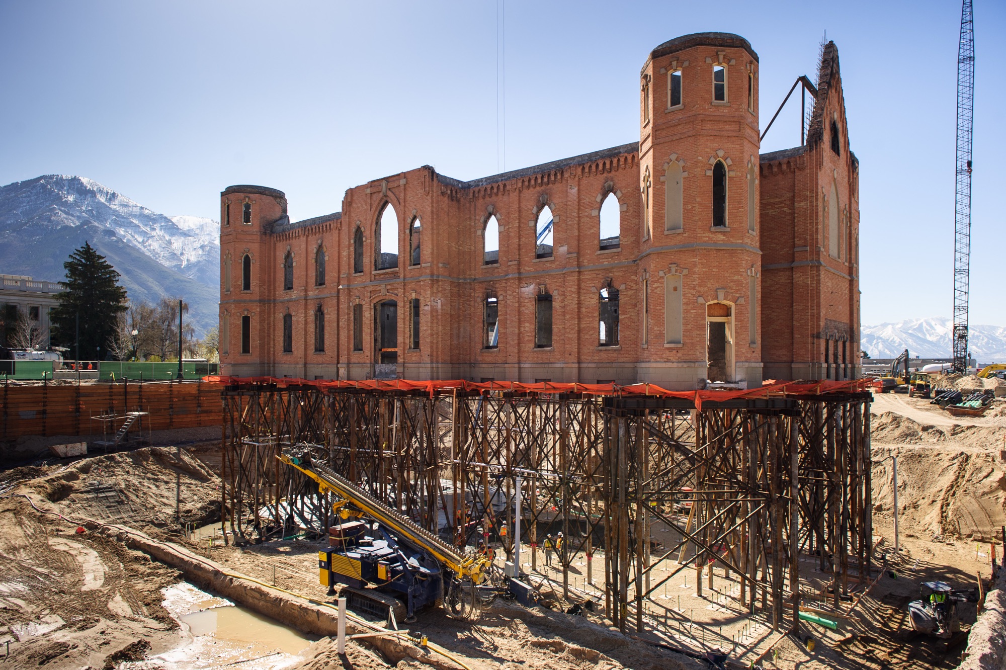 The historic Provo Tabernacle brick exterior held up by steel stilts during excavation with a large yellow excavator in the foreground.