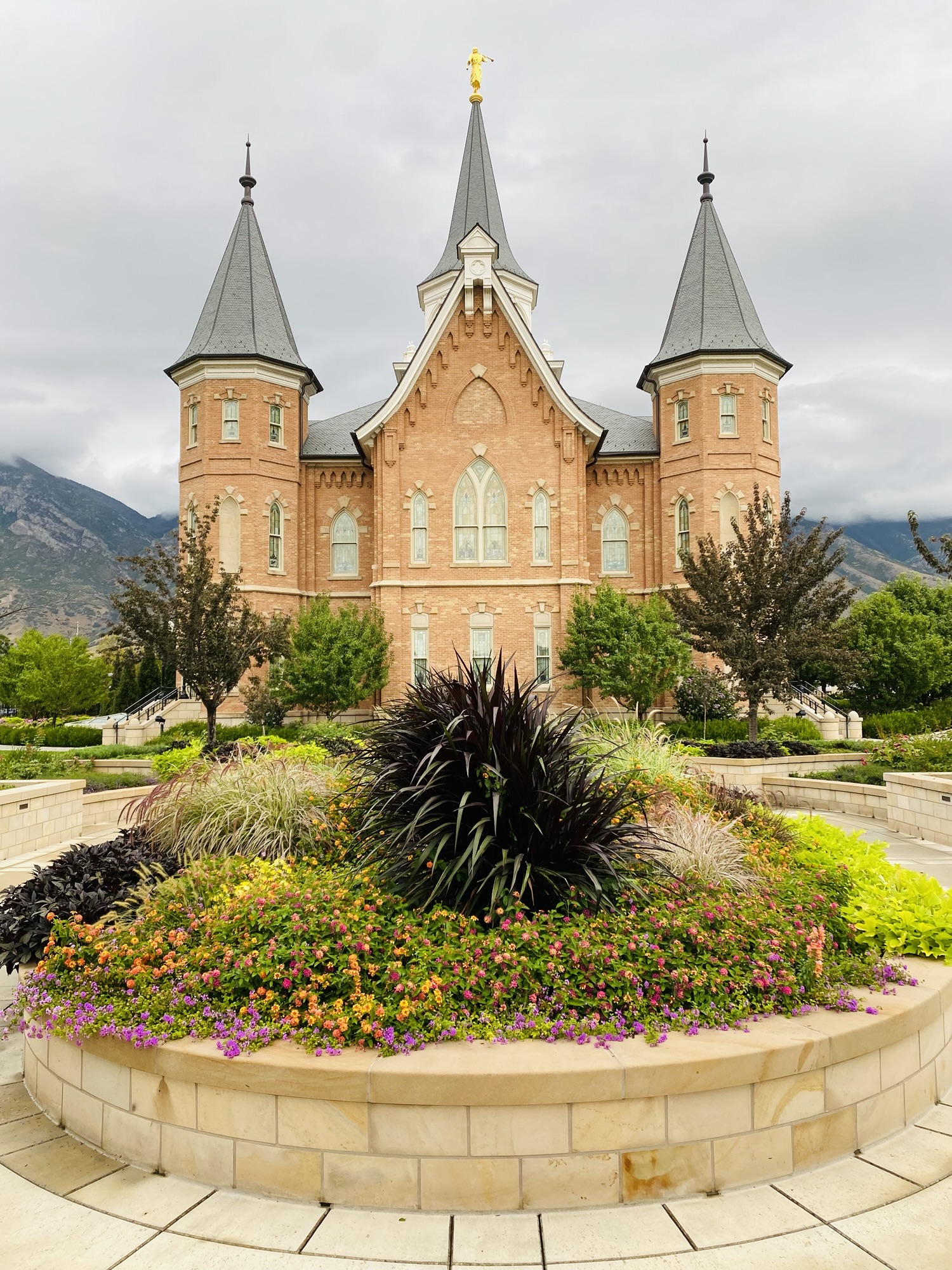 The finished Provo City Center Temple building viewed from the garden with colorful flowers and cloudy sky.