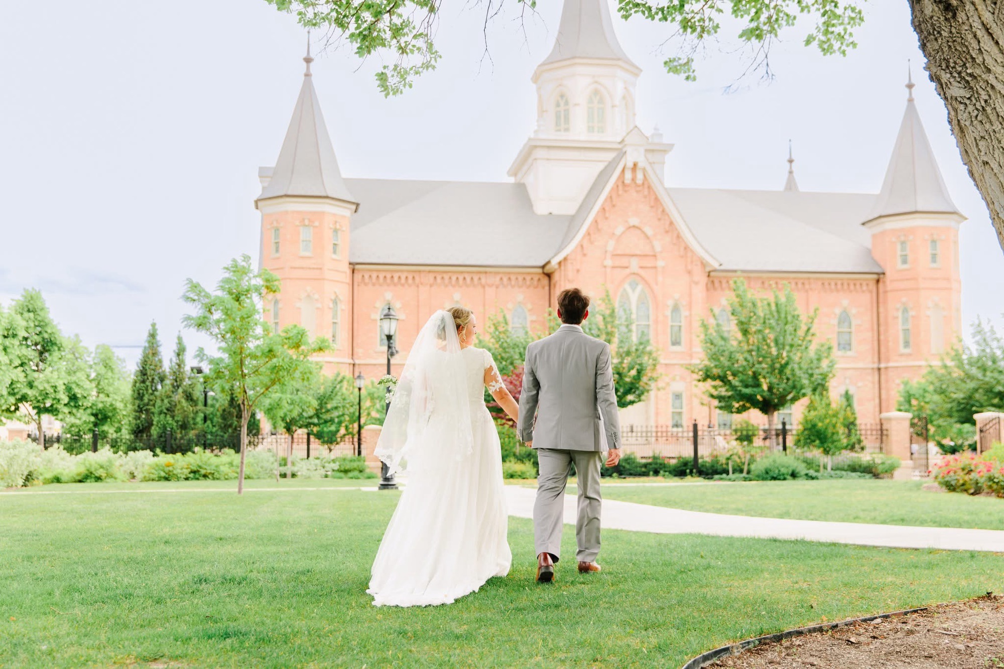Jared Harding Wilson in a wedding suit and his wife in a white wedding dress, seen from behind, holding hands and looking toward the Provo City Center Temple.