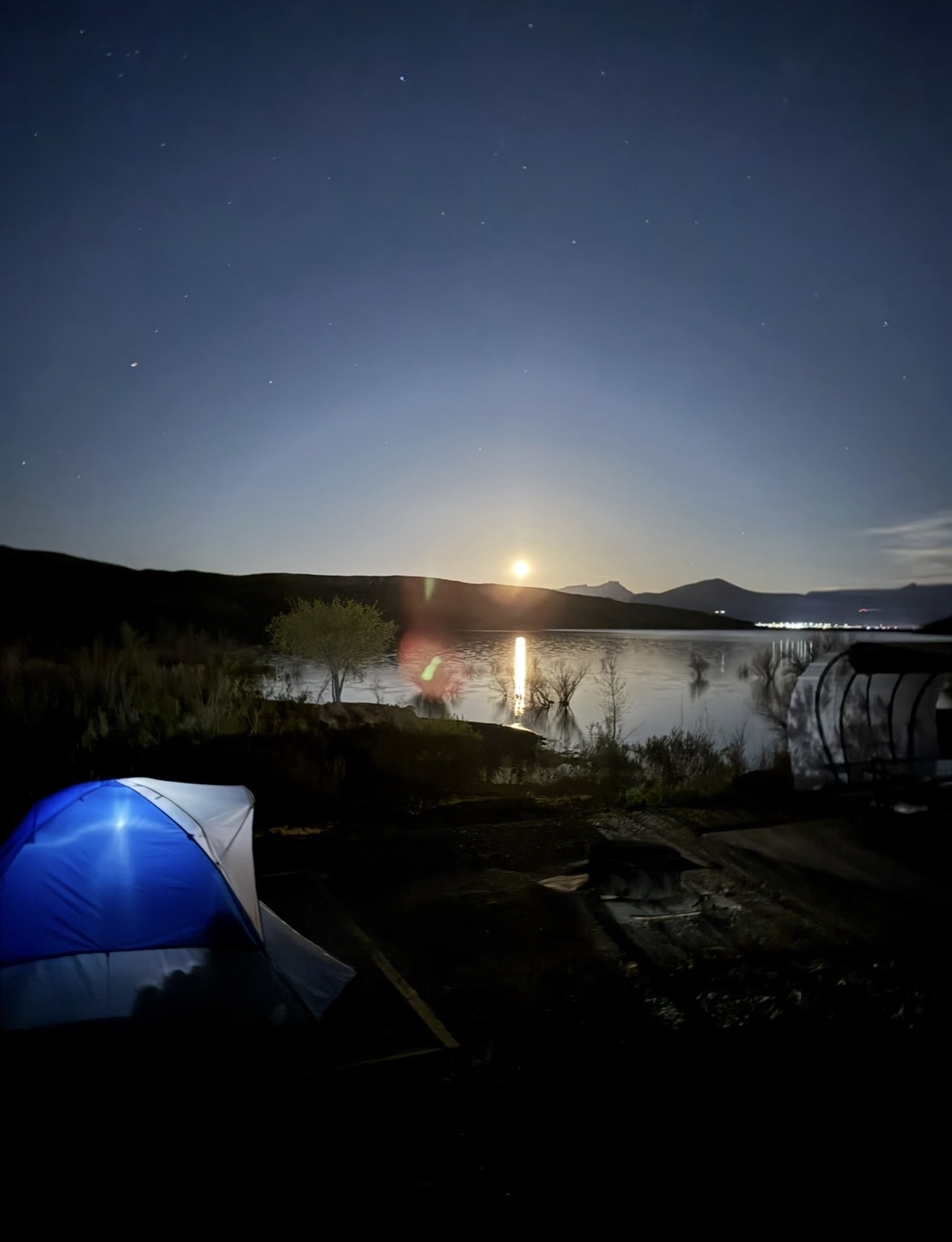 A blue camping tent pitched on the banks of a lake under a bright, full supermoon.
