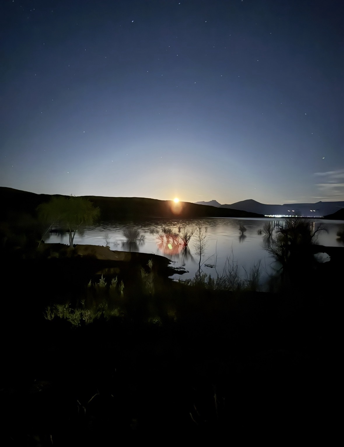 Vertical view of a glowing supermoon high in a dark, starry sky over a desert lake.