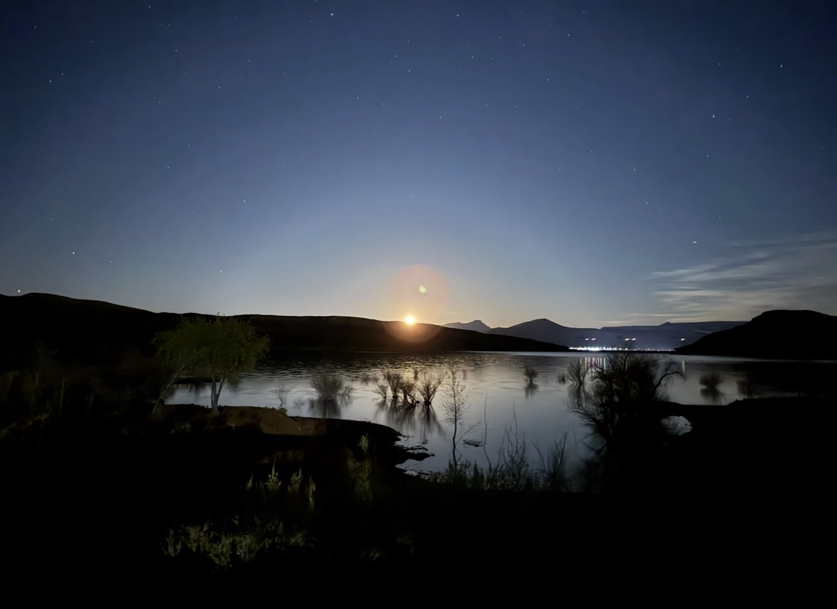 A bright supermoon rising over dark mountain silhouettes with stars visible above Quail Creek Reservoir beach.