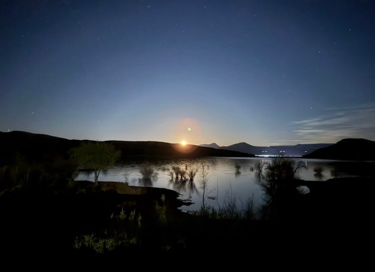 A bright supermoon rising over dark mountain silhouettes with stars visible above Quail Creek Reservoir beach.
