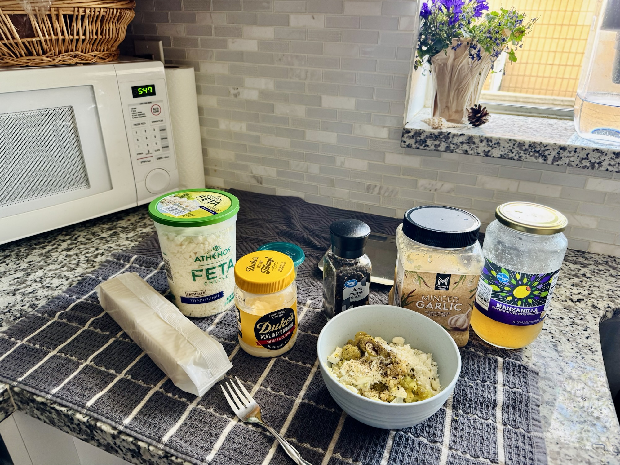 An overhead shot of the unmixed ingredients for Mediterranean egg salad including chopped eggs, mayo, feta, green olives, and pickles arranged in a bowl.