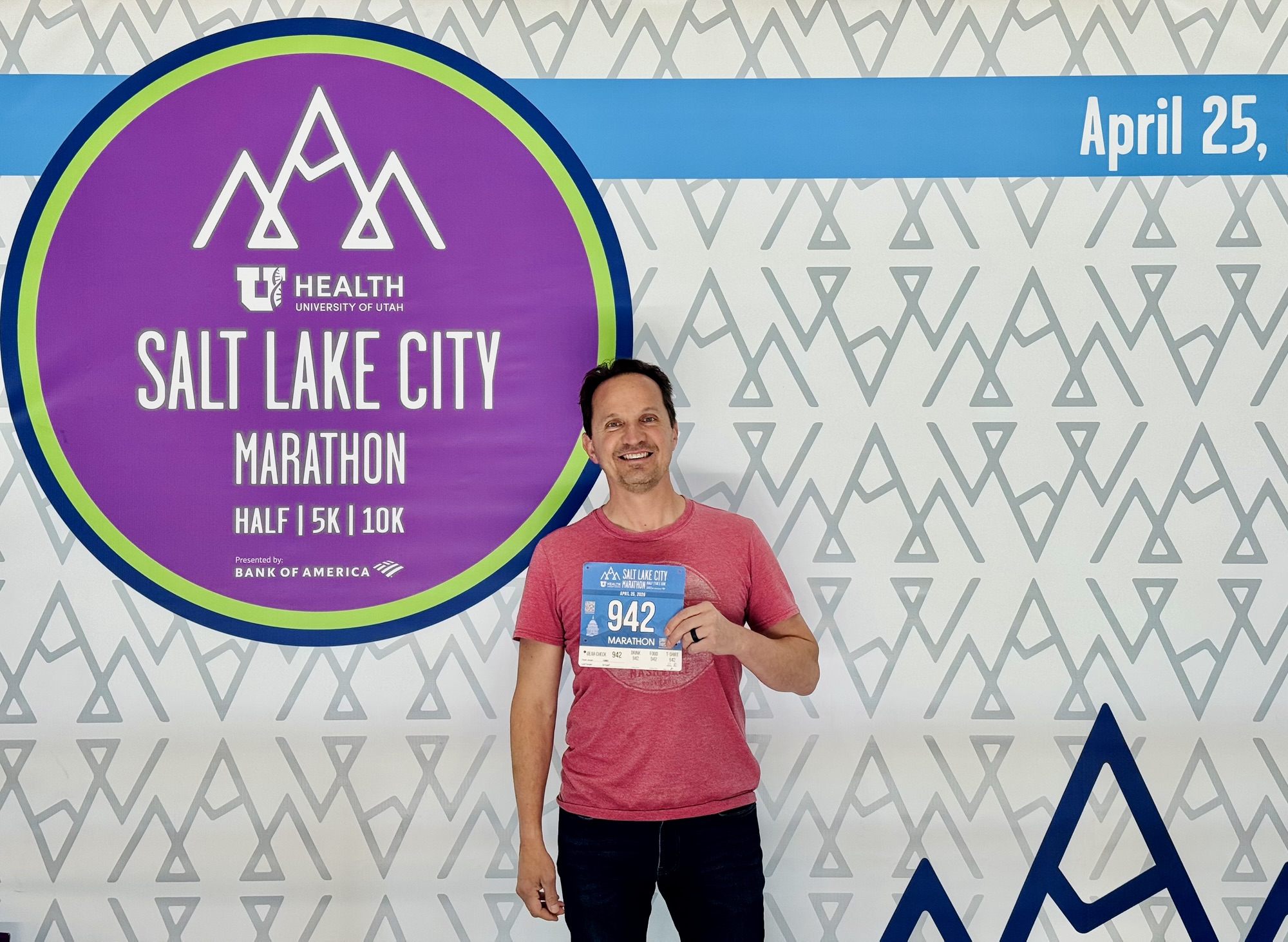 A photo of Jared Harding Wilson smiling and holding up a marathon race bib with the number 942 printed on it. They are standing in front of a large event sign that reads "Salt Lake Marathon." The atmosphere is bright and energetic, capturing the moment before the big race.