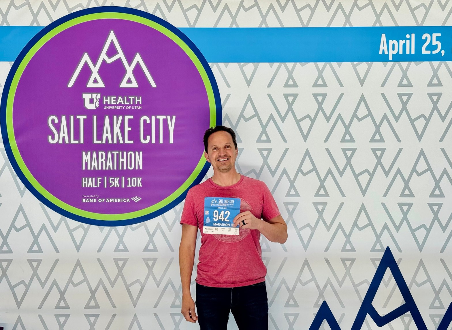 A photo of Jared Harding Wilson smiling and holding up a marathon race bib with the number 942 printed on it. They are standing in front of a large event sign that reads "Salt Lake Marathon." The atmosphere is bright and energetic, capturing the moment before the big race.