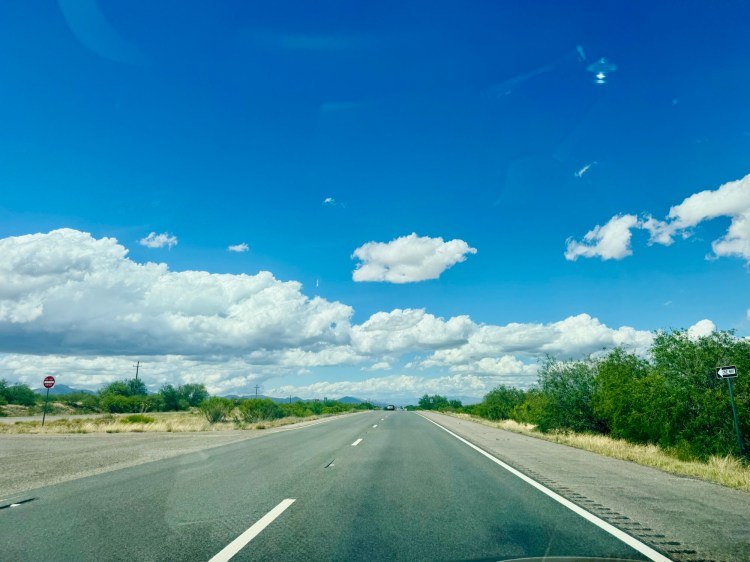 A scenic view of a highway heading toward Tucson, Arizona, under a bright blue sky with white puffy clouds and greenery on both sides, photographed by Jared Harding Wilson.