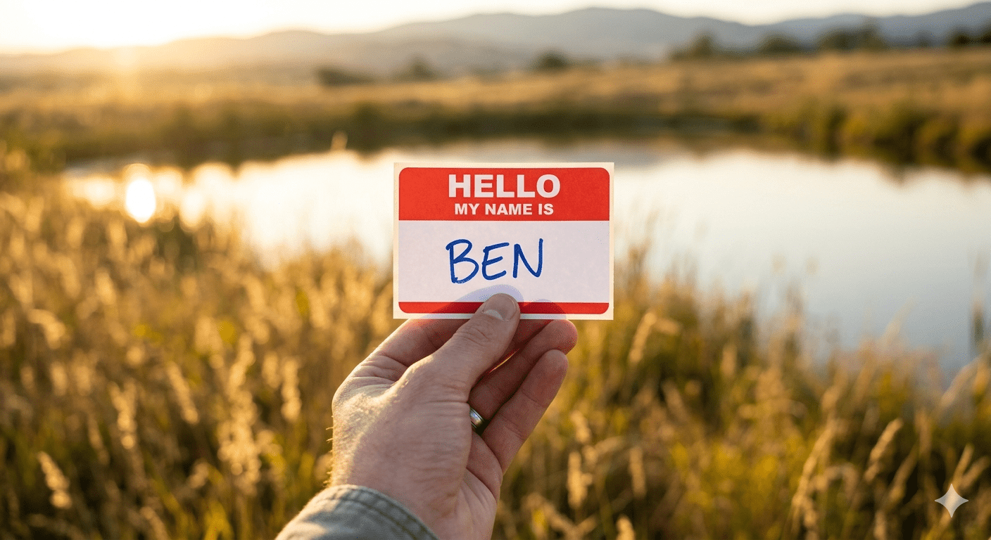 A close-up of a hand holding a red and white "Hello My Name Is" name tag with "Ben" handwritten in blue ink, set against a warm, reflective pond landscape at sunset. Photograph by Jared Harding Wilson.
